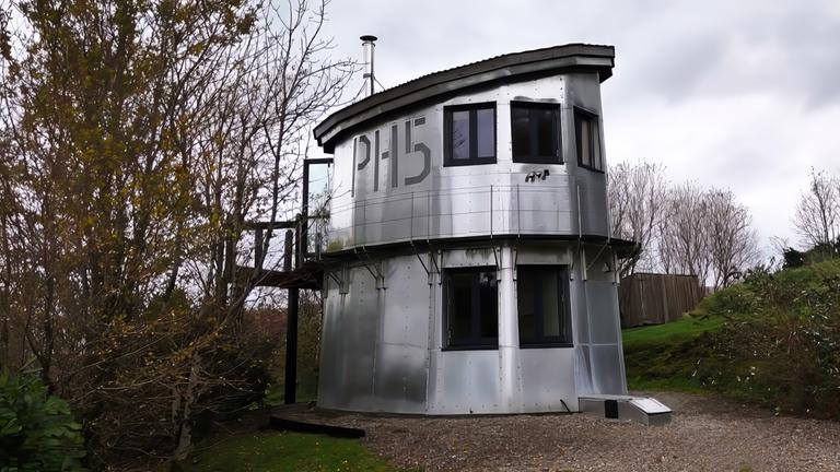 Ein silberner, zweistöckiger Wachturm mit großen Fenstern und der Nummer 'PH15' an der Seite steht an einem Weg im Grünen. Er ist umgeben von Bäumen und bewölktem Himmel