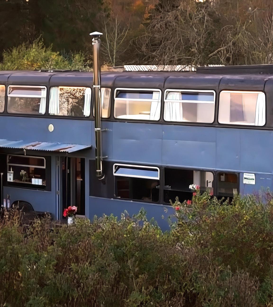 Ein blauer Doppeldeckerbus wurde in ein Haus umgebaut. Er steht im Grünen und hat mehrere Fenster, einem Schornstein und einen überdachten Eingang.