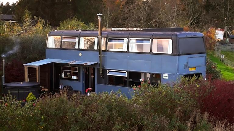 Ein blauer Doppeldeckerbus wurde in ein Haus umgebaut. Er steht im Grünen und hat mehrere Fenster, einem Schornstein und einen überdachten Eingang.