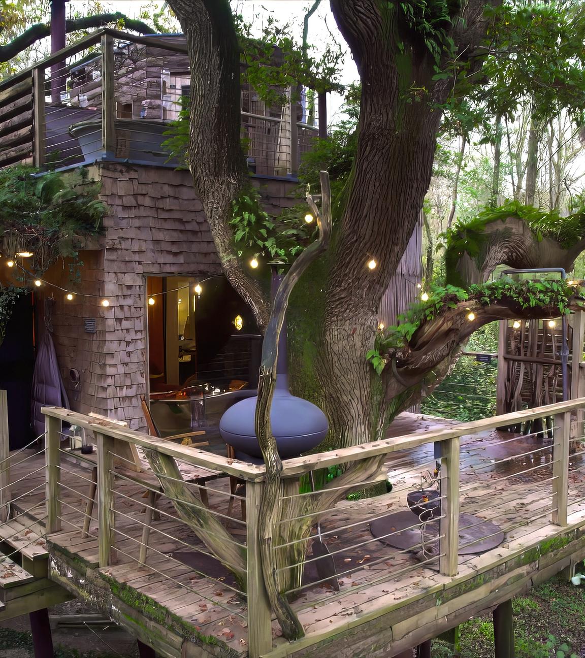 In einem großen Baum zwischen mehreren Ästen befindet sich ein Baumhaus mit Holzgeländer und Lichterkette, umgeben von weiteren Bäumen.