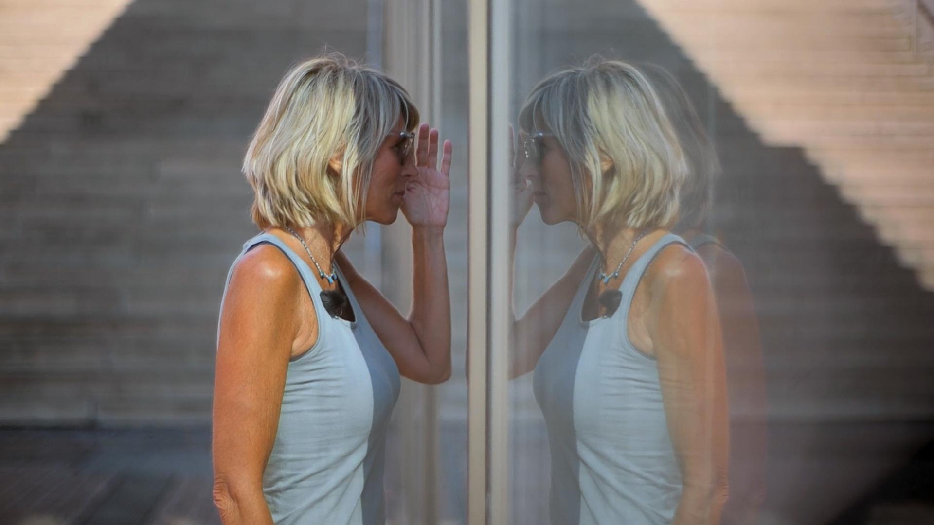 Steffi, eine sportlich schlanke Frau mit schulterlangen blonden Haaren steht an einer Glasscheibe und schaut hinein. Sie trägt eine Sonnenbrille und ein blaues Tank-Top. Neben ihr ist ihr Spiegelbild zu sehen.