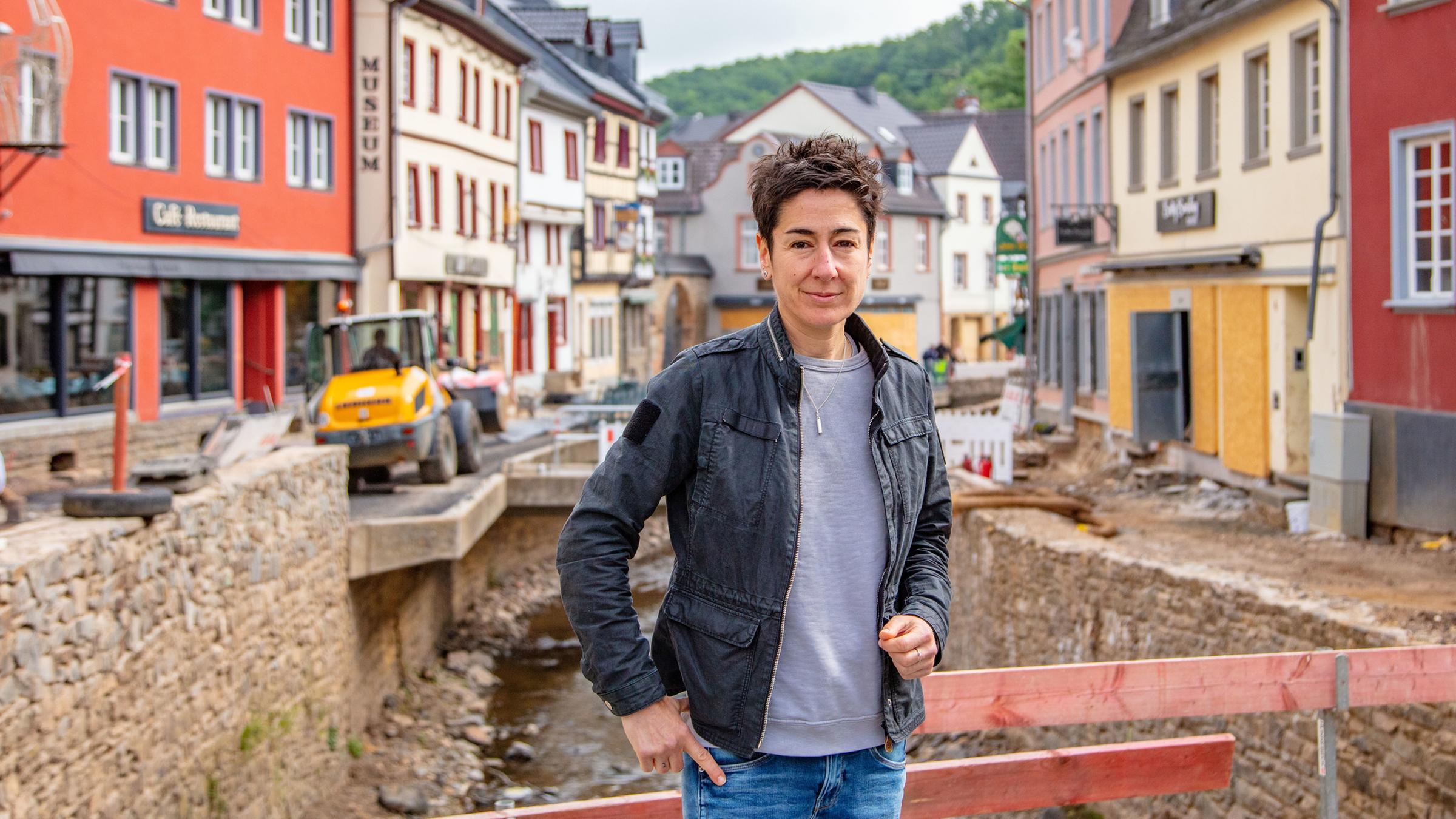 Im Bildvordergrund steht die ZDF-Reporterin Dunja Hayali auf einer Behelfsbrücke in der Fußgängerzone von Bad Münstereifel. Im Hintergrund sieht man Aufbauarbeiten am Flusslauf entlang der Erft. Sie fließt durch die historische Altstadt.