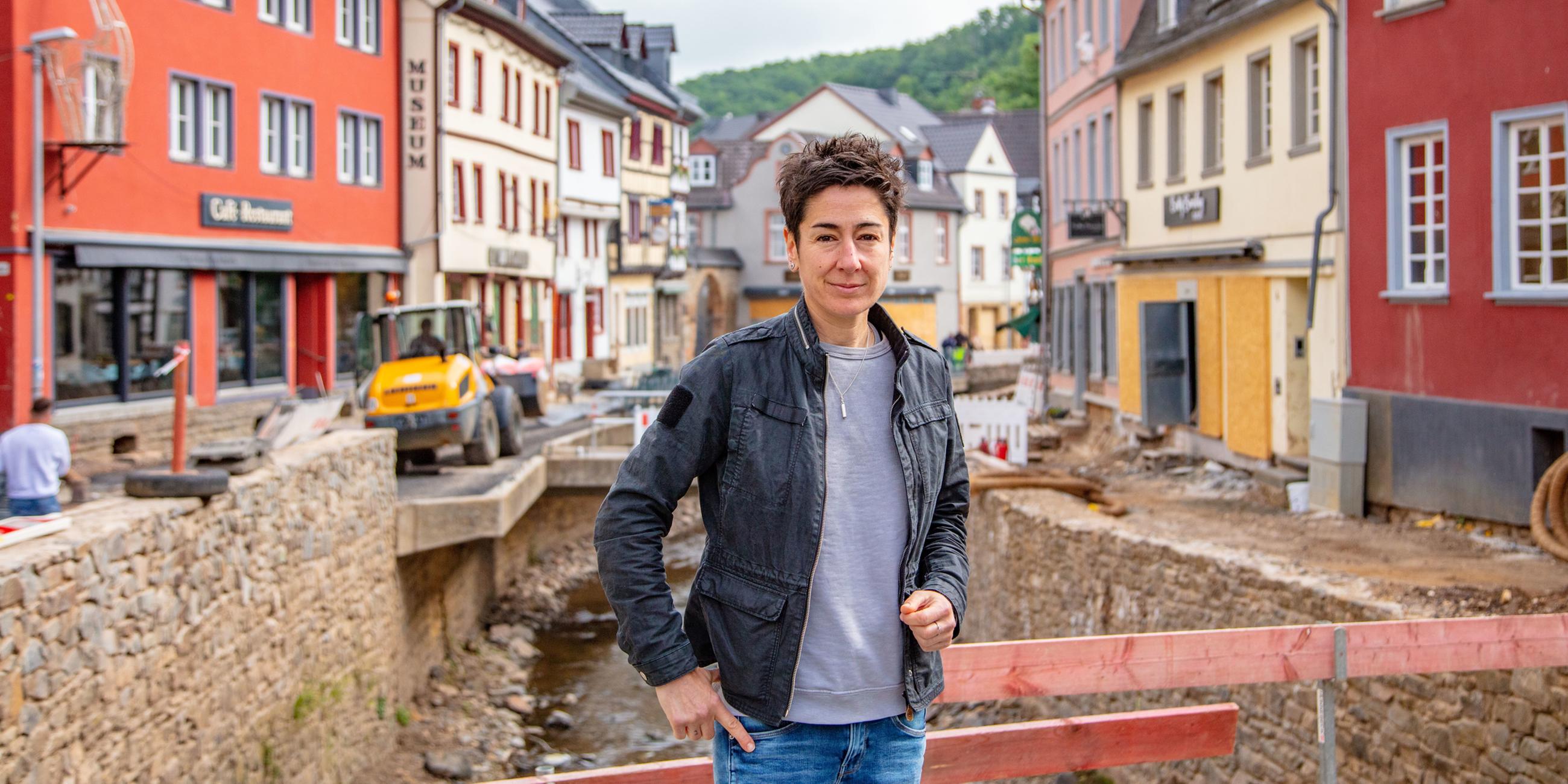 Im Bildvordergrund steht die ZDF-Reporterin Dunja Hayali auf einer Behelfsbrücke in der Fußgängerzone von Bad Münstereifel. Im Hintergrund sieht man Aufbauarbeiten am Flusslauf entlang der Erft. Sie fließt durch die historische Altstadt.