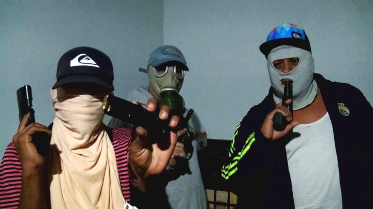 Drei Männer mit vermummten Gesichtern blicken in die Kamera. Sie haben Schusswaffen in der Hand, die sich an Mund und Schläfe halten.