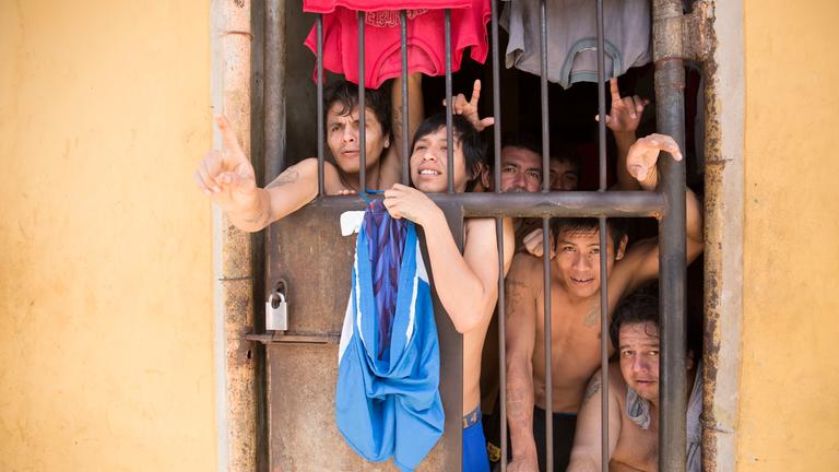 Fünf Männer mit nacktem Oberkörper zwängen sich hinter eine vergitterte Türe und schauen nach draußen.