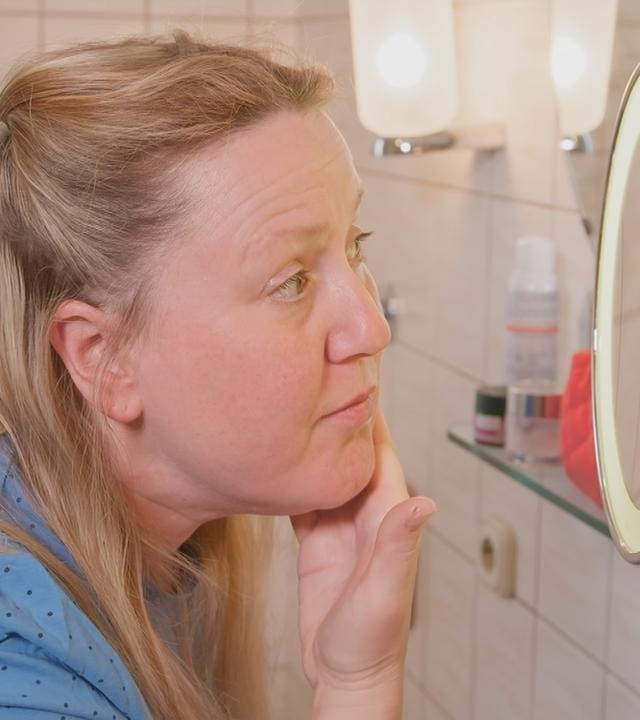 Frau blickt in den Spiegel und betrachtet ihre Haut.