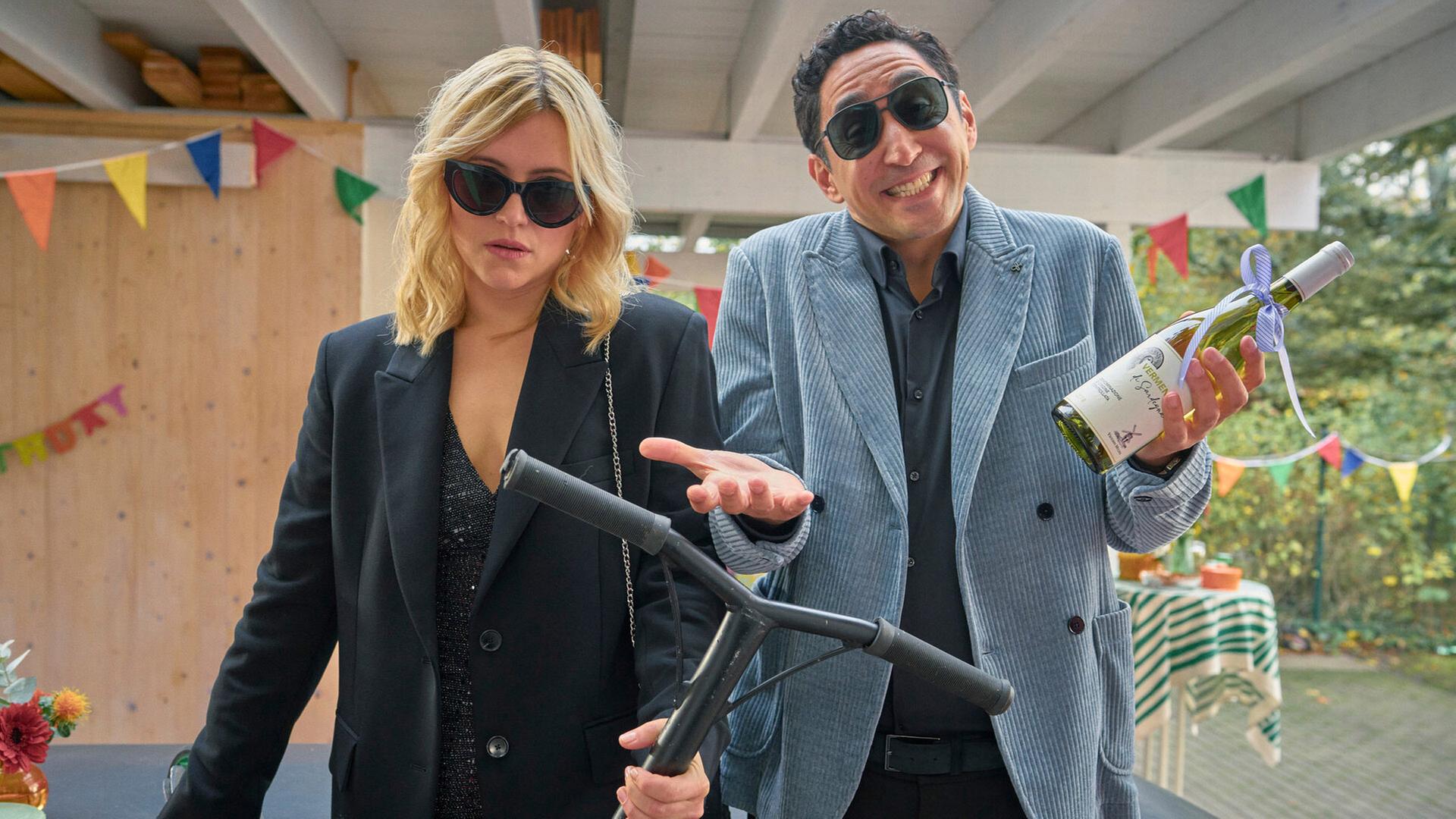 "Die Kinderschwindlerin": Nina (Laura Storz) und ihr Freund Sammy (Ekrem "Eko Fresh" Bora) stehen auf einer bunt geschmückten Veranda. Nina schaut genervt, während Sammy lacht.