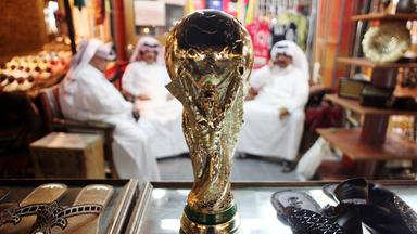 Im Vordergrund Nachbildung des FIFA WM-Pokals zwischen zwei Paar Schuhen. Im Hintergrund arabische Männer im Halbkreis sitzend.