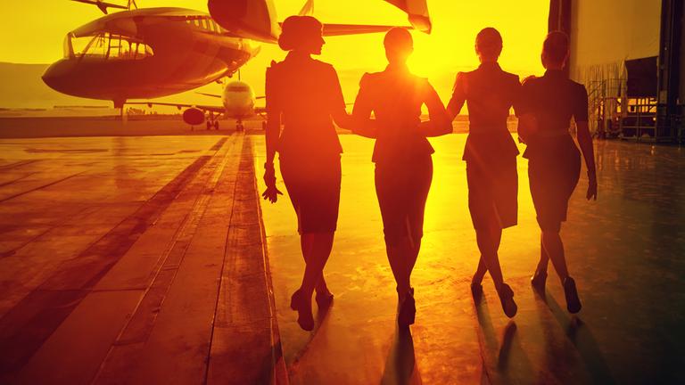 Eine Gruppe Flugbegleiterinnen von hinten vor einem Flugzeug in den Sonnenuntergang.