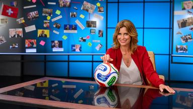 Moderatorin Valerie Niehaus sitzt am Schreibtisch im Studio, hält einen Fußball in der Hand und blickt in die Kamera.