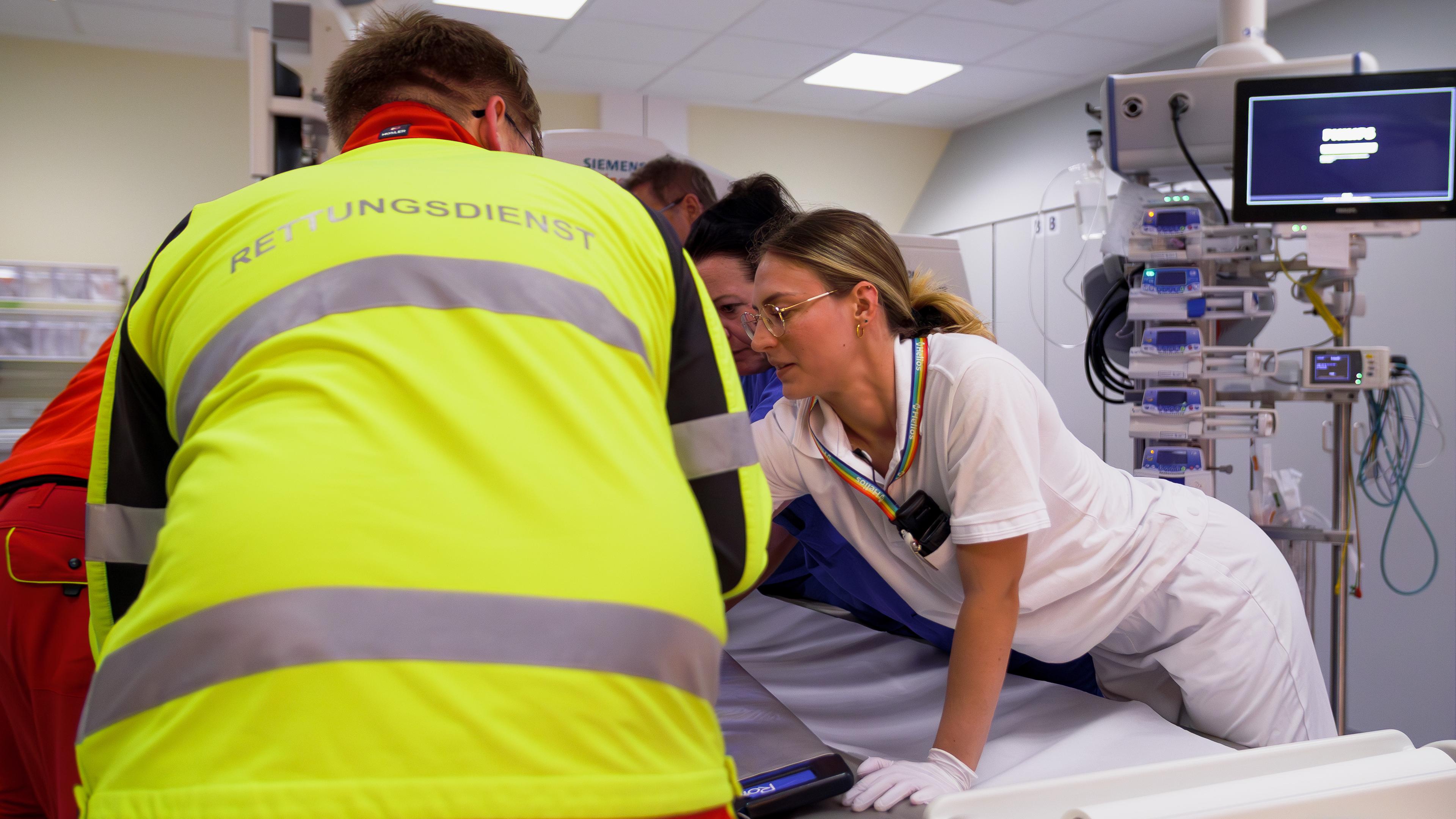 Ein Patient liegt auf einem Behandlungstisch. Im Vordergrund steht ein Rettungssanitäter mit neon-gelber Weste. Daneben steht Assistenzärztin Jule, die sich um den Patienten kümmert.