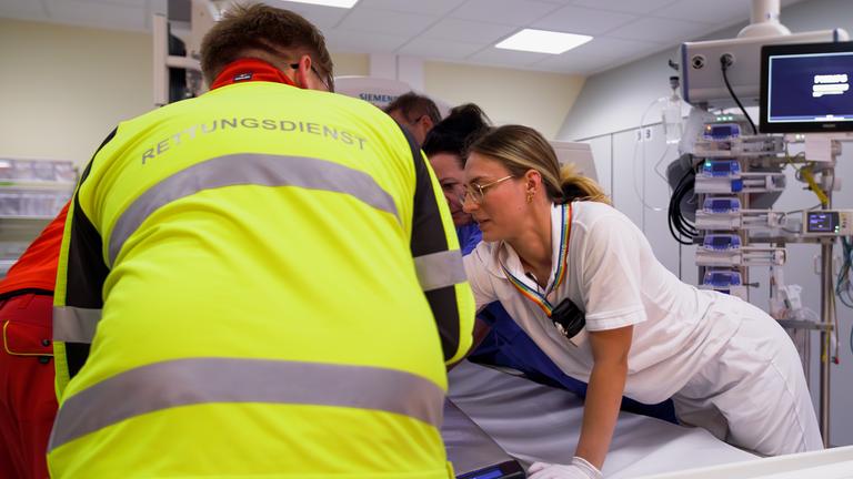 Ein Patient liegt auf einem Behandlungstisch. Im Vordergrund steht ein Rettungssanitäter mit neon-gelber Weste. Daneben steht Assistenzärztin Jule, die sich um den Patienten kümmert.