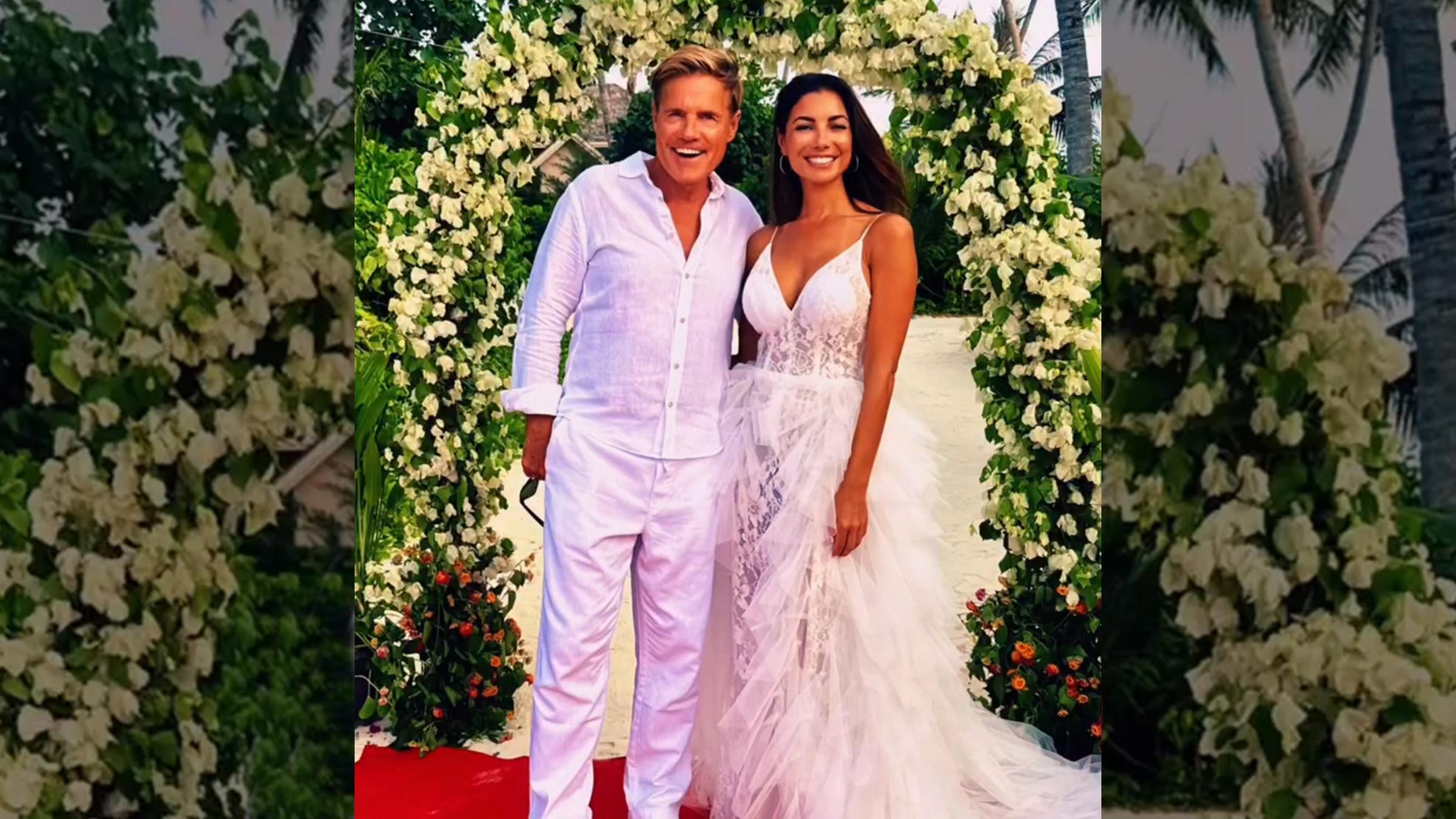 Dieter Bohlen (links) neben seiner neuen Ehefrau Carina Walz (rechts), beide tragen weiß und stehen vor einem mit Blumen geschmückten Torbogen. 