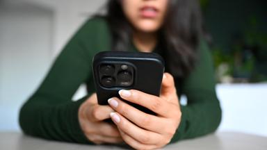 Eine junge Frau sitzt mit ihrem Handy an einem Tisch und blickt auf das Display