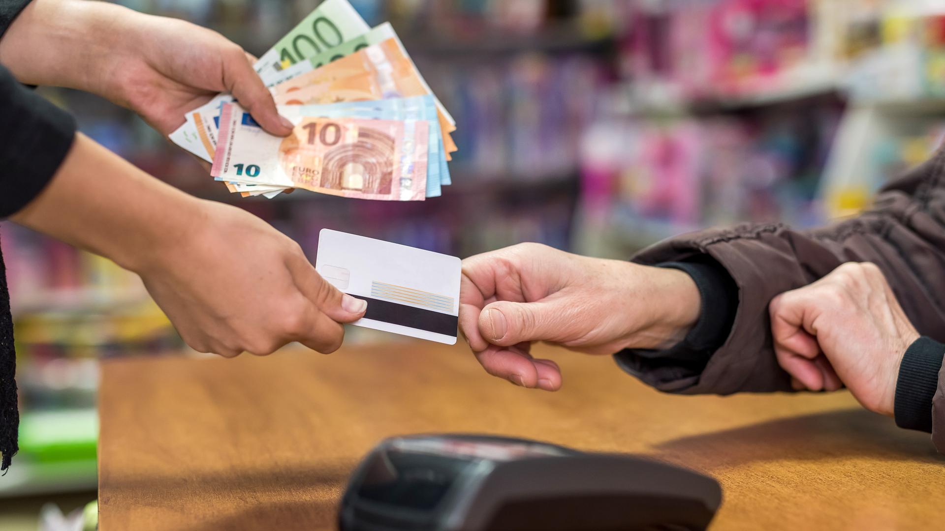 Eine Person hält in der einen Hand Geldscheine und überreicht mit der anderen Hand einer Person eine Kreditkarte.
