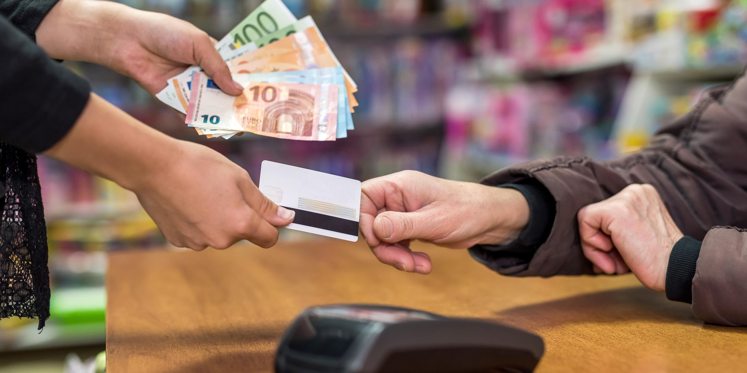 Eine Person hält in der einen Hand Geldscheine und überreicht mit der anderen Hand einer Person eine Kreditkarte.
