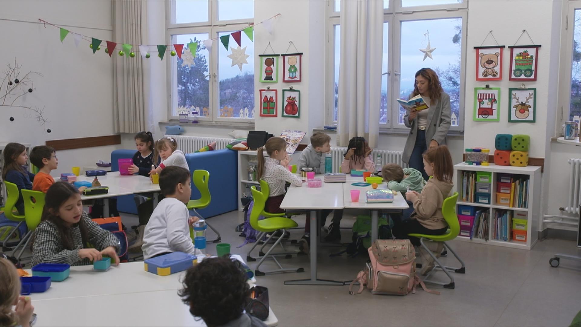 Auf dem BIld sieht man eine Lehrerin mit ihren Schüler*innen im Klassenzimmer.