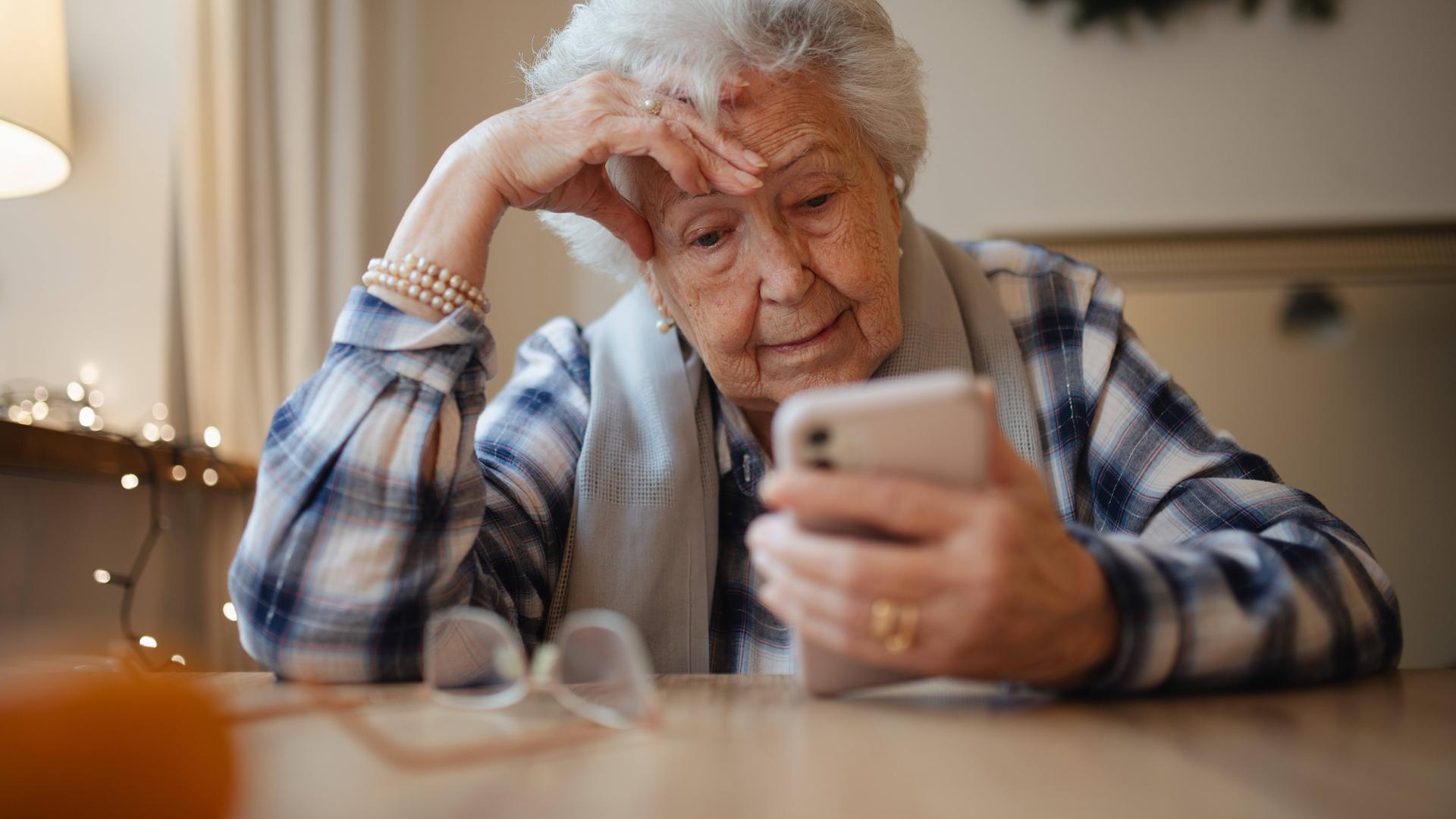 Eine Seniorin schaut auf ein Smartphone und stützt ihren Kopf auf der rechten Hand ab.