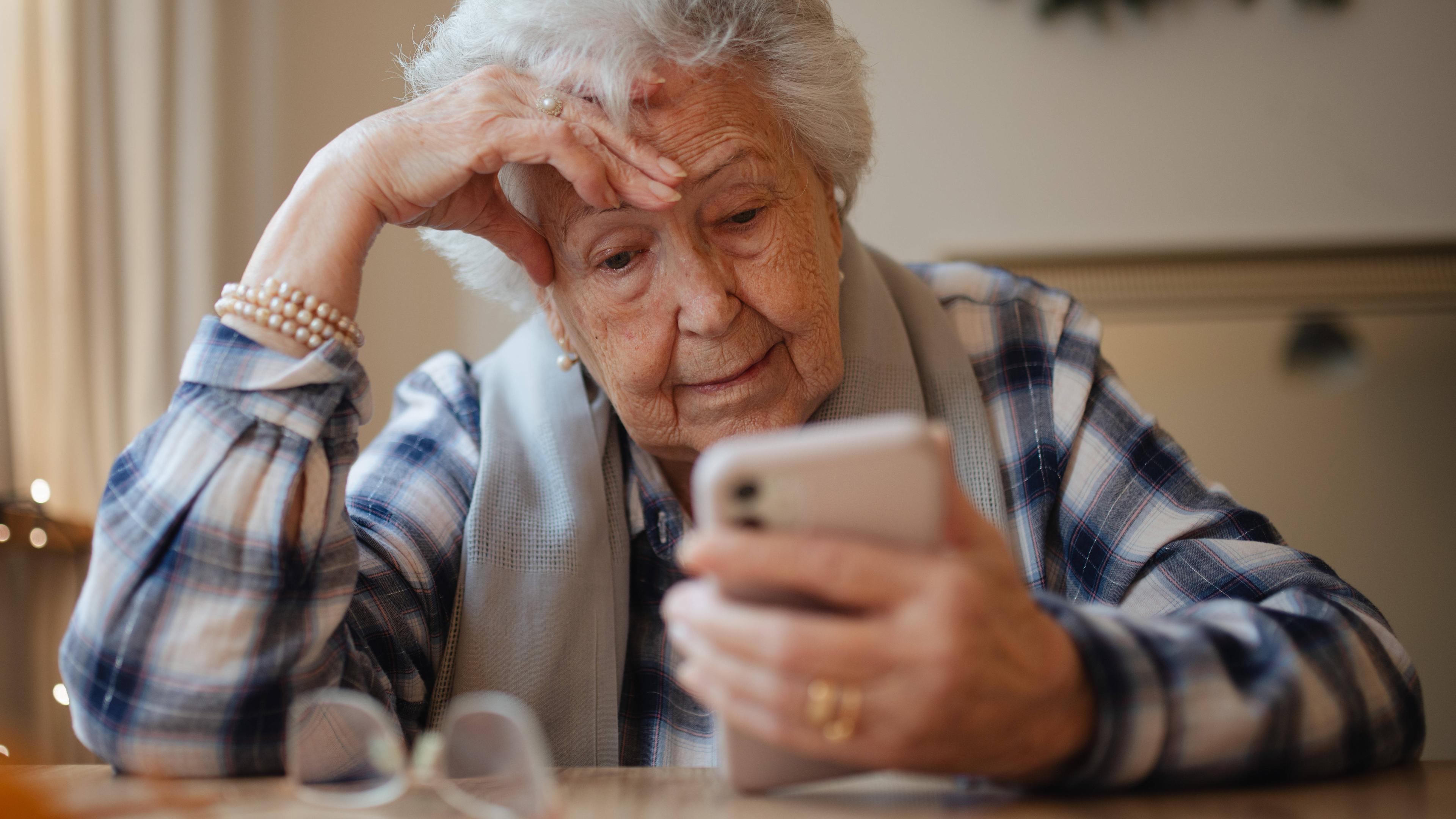 Eine Seniorin schaut auf ein Smartphone und stützt ihren Kopf auf der rechten Hand ab.