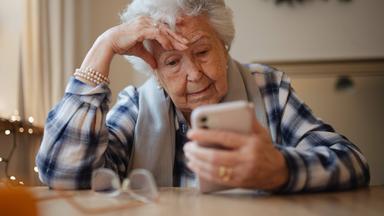 Eine Seniorin schaut auf ein Smartphone und stützt ihren Kopf auf der rechten Hand ab.