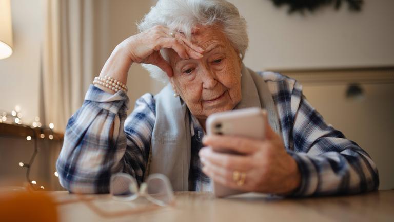 Eine Seniorin schaut auf ein Smartphone und stützt ihren Kopf auf der rechten Hand ab.