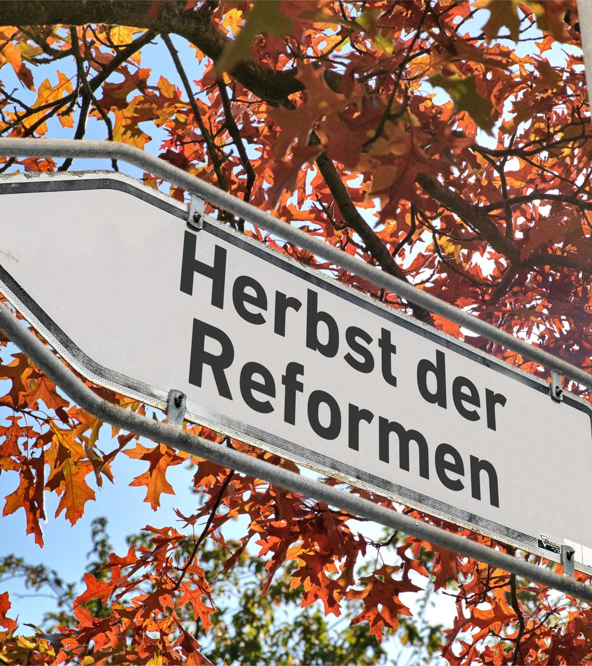 Ein weißes Hinweisschild mit abgeblaetterter Farbe am Rand und der Aufschrift "Herbst der Reformen" an einem Laternenmast als Wegweiser nach links vor einem Baum mit herbstlich verfärbten Blättern.