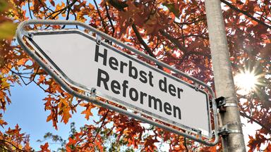 Ein weißes Hinweisschild mit abgeblaetterter Farbe am Rand und der Aufschrift "Herbst der Reformen" an einem Laternenmast als Wegweiser nach links vor einem Baum mit herbstlich verfärbten Blättern.