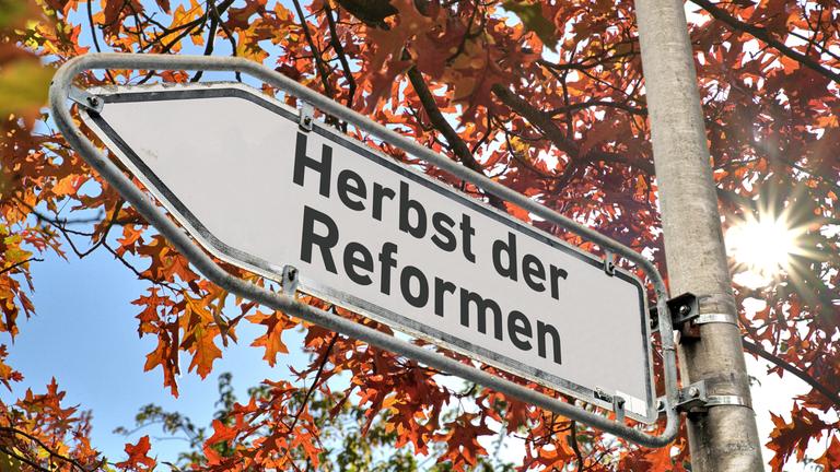 Ein weißes Hinweisschild mit abgeblaetterter Farbe am Rand und der Aufschrift "Herbst der Reformen" an einem Laternenmast als Wegweiser nach links vor einem Baum mit herbstlich verfärbten Blättern.