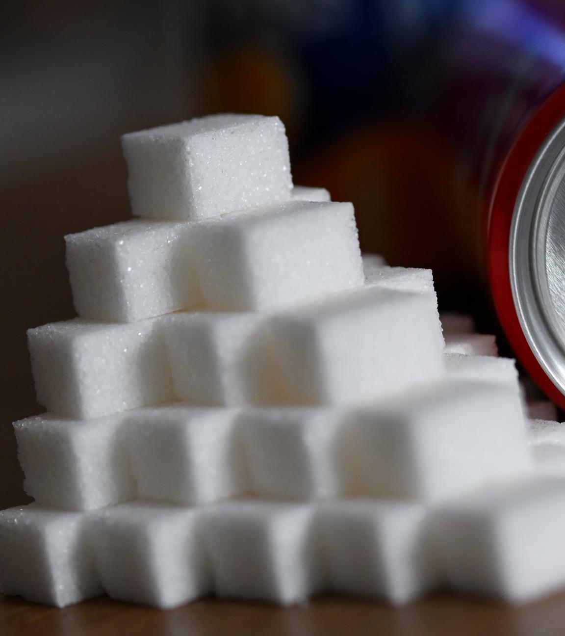 ine Getränkedose mit einem Softdrink und zahlreiche Zuckerwürfel liegen auf einem Tisch.