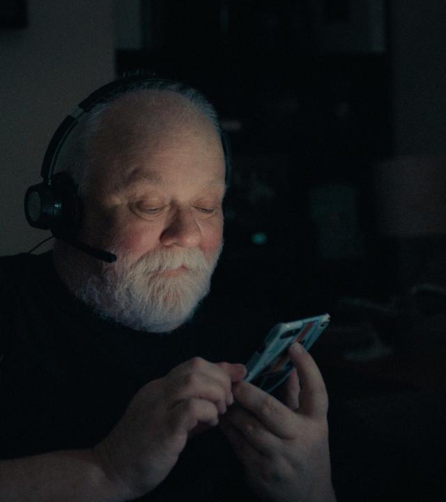 Ein älterer Mann mit glatze und grauem Bart trägt ein Headset und schaut lächelnd auf ein Handydisplay, das ihn im Dunkeln anleuchtet.