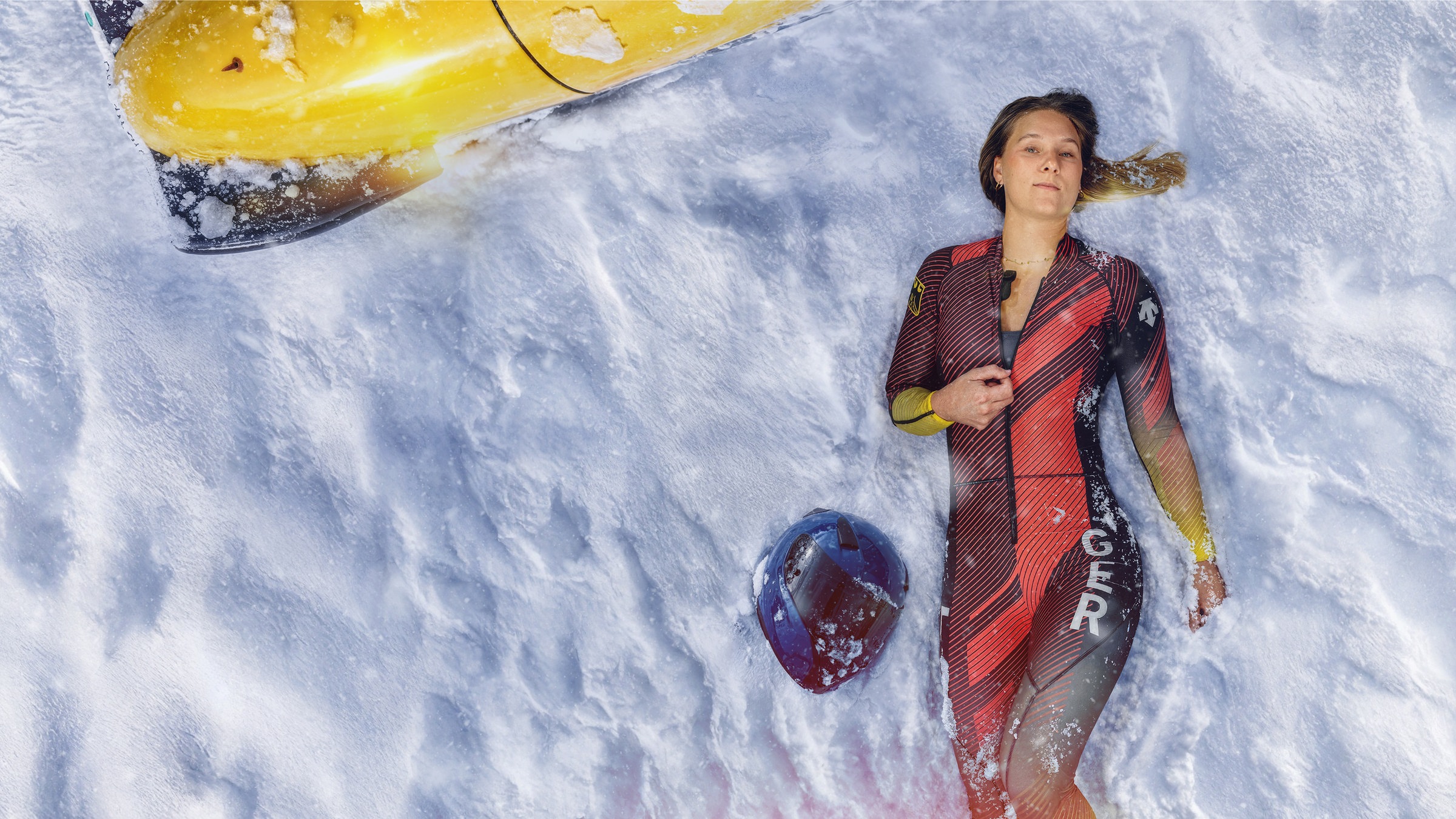 Lisa Buckwitz liegt, aus der Vogelperspektive fotografiert, im Schnee und trägt ihren Sportanzug, neben ihr liegt ihr Sporthelm im Schnee. Sie deutet mit der Hand an, den Reißverschluss ihres Anzuges zu öffnen.