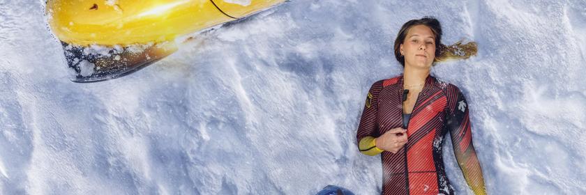 Lisa Buckwitz liegt, aus der Vogelperspektive fotografiert, im Schnee und trägt ihren Sportanzug, neben ihr liegt ihr Sporthelm im Schnee. Sie deutet mit der Hand an, den Reißverschluss ihres Anzuges zu öffnen.