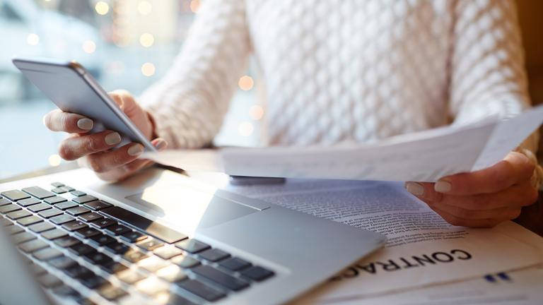Eine Person in weißem Pullover sitzt an einem Tisch. Sie hält ein Smartphone in der einen und ein Papier in der anderen Hand. An der linken Seite steht ein Laptop.