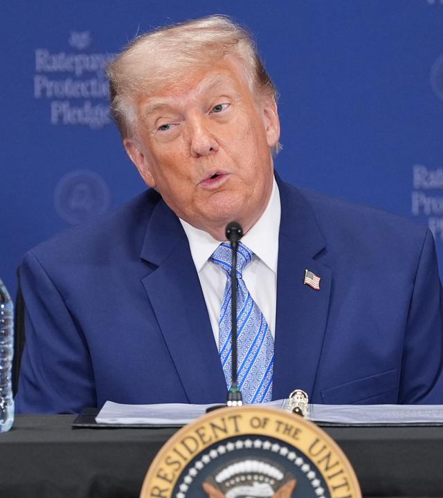 Donald Trump sitzt an einem Rednerpult und spricht in ein Mikrofon.