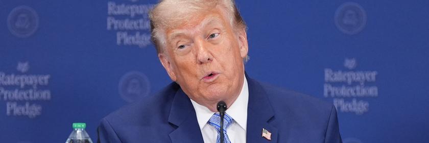 Donald Trump sitzt an einem Rednerpult und spricht in ein Mikrofon.