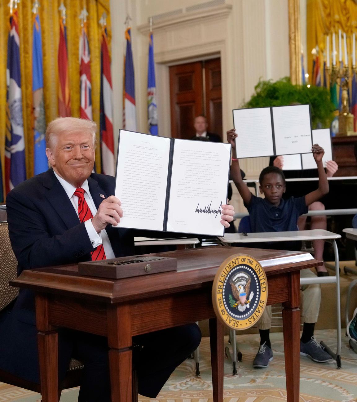 Donald Trump hält ein unterzeichnetes Dekret hoch, während junge Menschen im Hintergrund Kopien davon halten.