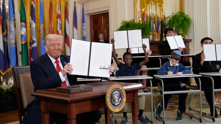 Donald Trump hält ein unterzeichnetes Dekret hoch, während junge Menschen im Hintergrund Kopien davon halten.