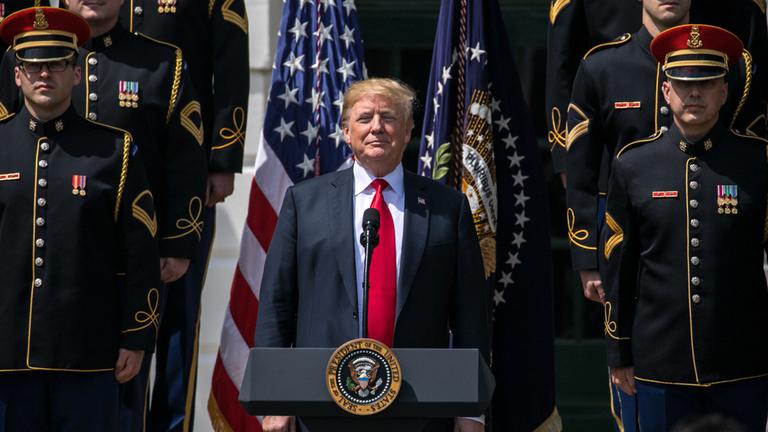 Donald Trump vor einer Amerika-Flagge und Soldaten, steht hinter einem Rednerpult.
