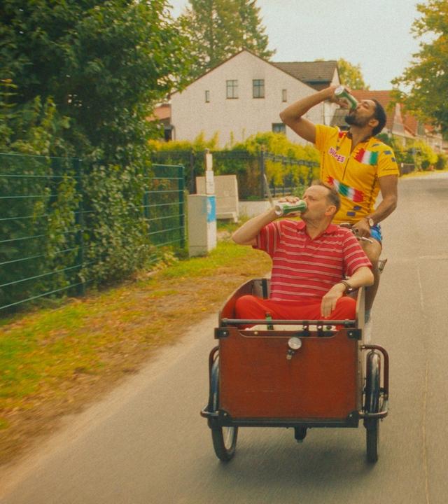 Doppelhaushälfte: Zwei Männer fahren auf einer Straße mit einem Lastenrad durch ein Wohngebiet und trinken dabei Bier. Andi (Milan Peschel) sitzt vorne im Lastenkorb, Theo (Benito Bause) auf dem Fahrrad. 