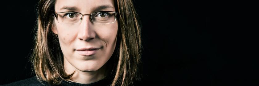 Eine lächelnde Frau mit langen Haaren und Brille schaut den Betrachter direkt an.