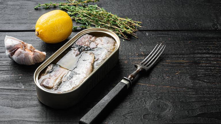 Eine geöffnete Dose mit eingelegtem Fisch steht auf einem Tisch, daneben eine Knoblauchknolle, eine Zitrone, Kräuter und eine Gabel.