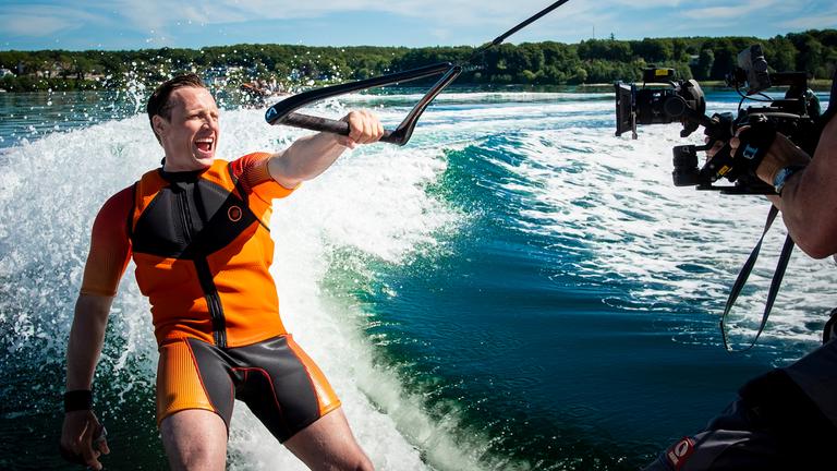 Schauspieler Patrick Kalupa fährt bei Dreharbeiten Wasserski und hält sich mit nur einer Hand fest.