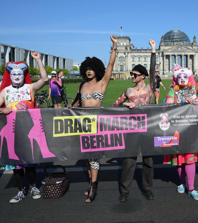 Sechs Drags* stehen vor dem Reichstag in Berlin. Sie halten ein Banner auf dem steht: Drag* March Berlin.