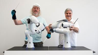 Frank H. P. Fitzek, Sprecher des Exzellenzcluster CeTI (Centre for Tactile Internet with Human-in-the-Loop) der Technischen Universität Dresden, steht mit Datenhandschuhen neben Markus Rindt, Intendant der Dresdner Sinfoniker, an zwei kollaborativen Robotern, die ein Orchester dirigieren sollen
