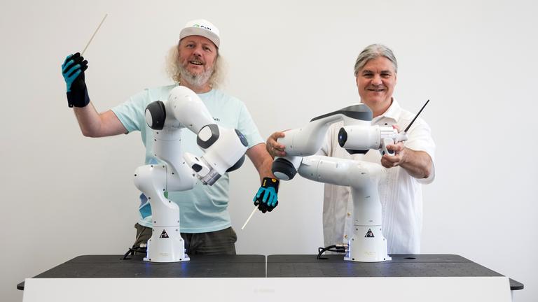 Frank H. P. Fitzek, Sprecher des Exzellenzcluster CeTI (Centre for Tactile Internet with Human-in-the-Loop) der Technischen Universität Dresden, steht mit Datenhandschuhen neben Markus Rindt, Intendant der Dresdner Sinfoniker, an zwei kollaborativen Robotern, die ein Orchester dirigieren sollen
