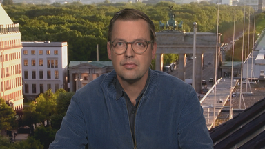 Christan Rode aus dem Berliner Studio zugeschalten. Er trägt eine Petrol-farbene Jacke und eine runde Brille.