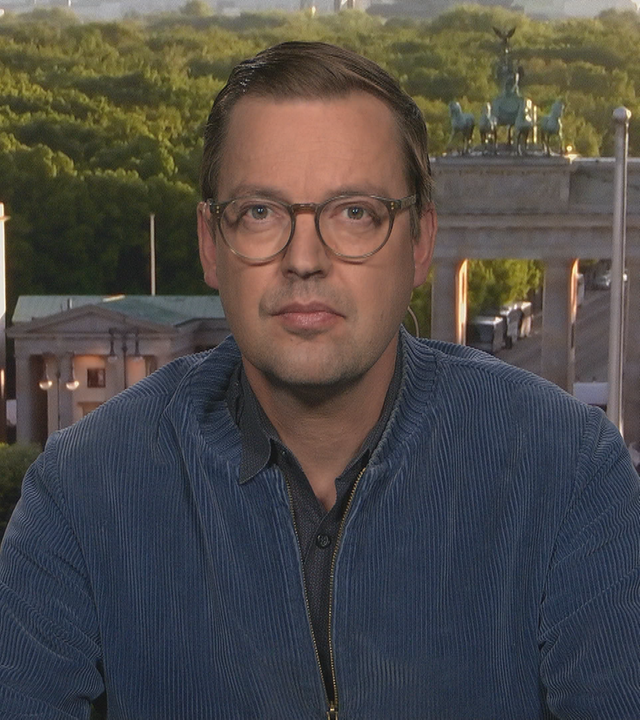 Christan Rode aus dem Berliner Studio zugeschalten. Er trägt eine Petrol-farbene Jacke und eine runde Brille.