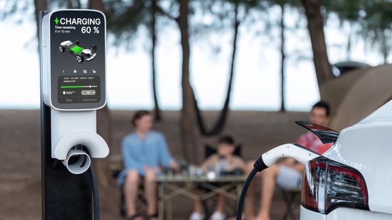 Eine Familie sitzt an einem Campingtisch. Im Vordergrund ist ein E-Auto an einer Ladesäule angeschlossen.