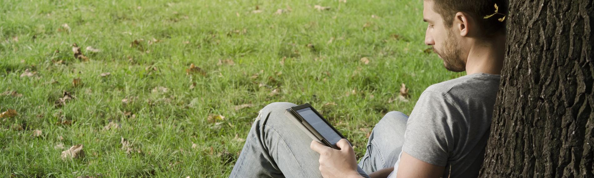 Ein Mann sitzt an einen Baumstamm lehnend auf einer Wiese und liest ein E-Book.