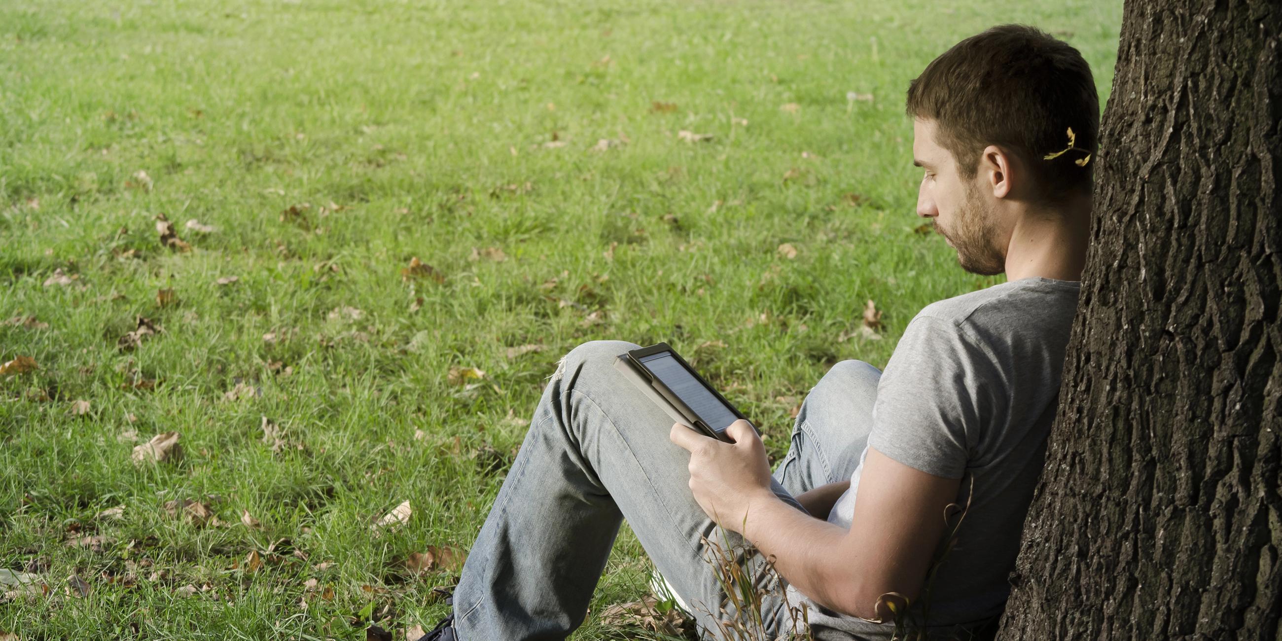 Ein Mann sitzt an einen Baumstamm lehnend auf einer Wiese und liest ein E-Book.